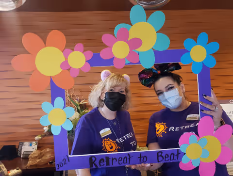Two women wearing purple Retreat at Sunny Vista t-shirts and face masks pose behind a colorful flower-decorated frame that reads 'Retreat to Beat 2021'. They are indoors with a wooden wall background.