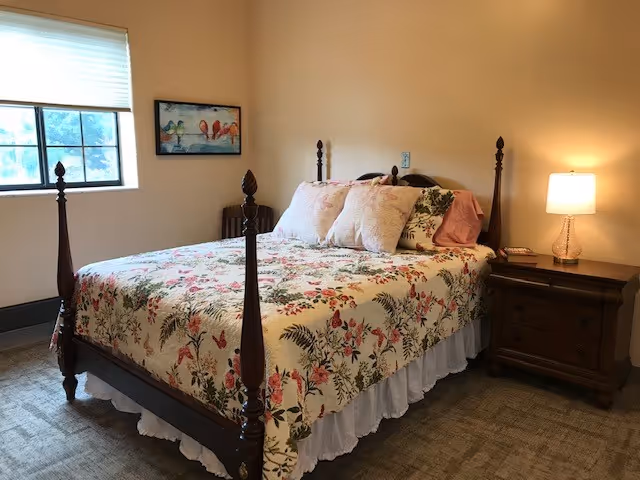A cozy bedroom with a wooden four-poster bed covered in a floral quilt and multiple pillows. There is a wooden nightstand with a lamp and a phone on it. A window with blinds lets in natural light, and a colorful bird painting hangs on the wall.