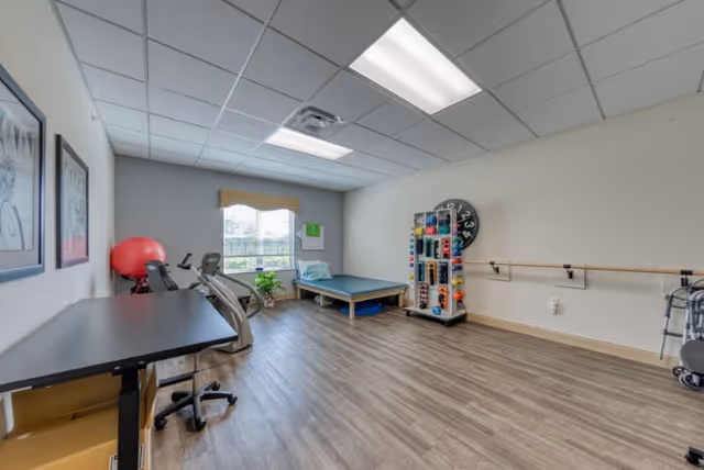 A rehabilitation or physical therapy room with exercise equipment including a stationary bike, a large red exercise ball, a padded therapy table, a rack with various weights, and a walker. The room has wood-style flooring, a window with a valance, and framed pictures on the wall.