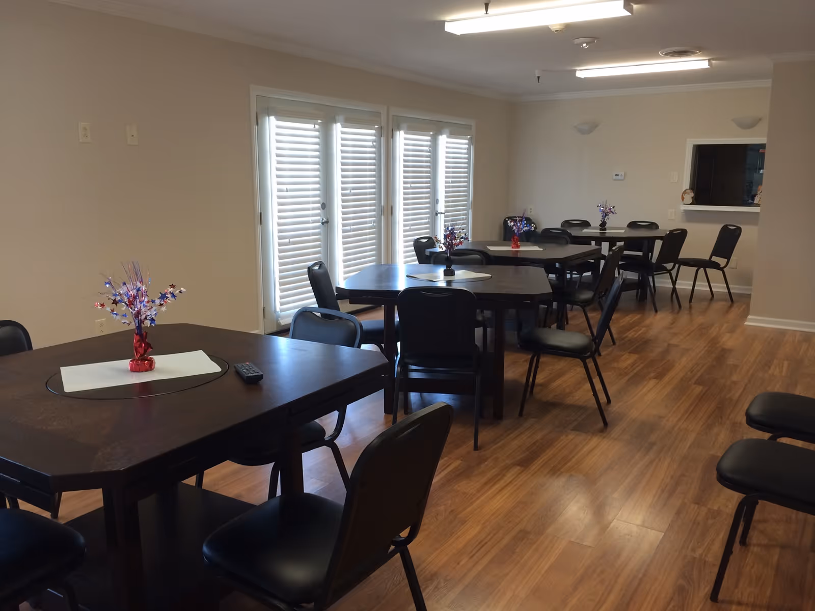 Interior view of a dining room with multiple dark wooden tables and black chairs arranged neatly. Each table has a small decorative centerpiece with red, white, and blue star-shaped decorations. The room has wooden flooring, beige walls, and large windows with white blinds letting in natural light. There is a pass-through window to another room on the right wall.