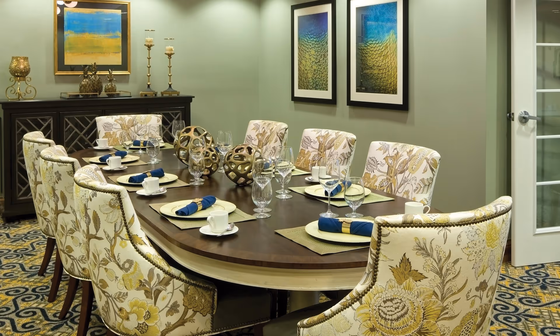 A formal dining room with a large wooden oval table set for eight people. The table is decorated with blue napkins, white plates, glassware, and decorative gold spheres. The chairs around the table have floral upholstery. The room has green walls adorned with framed artwork and a sideboard with candles and decorative items. A glass-paneled door is visible on the right.