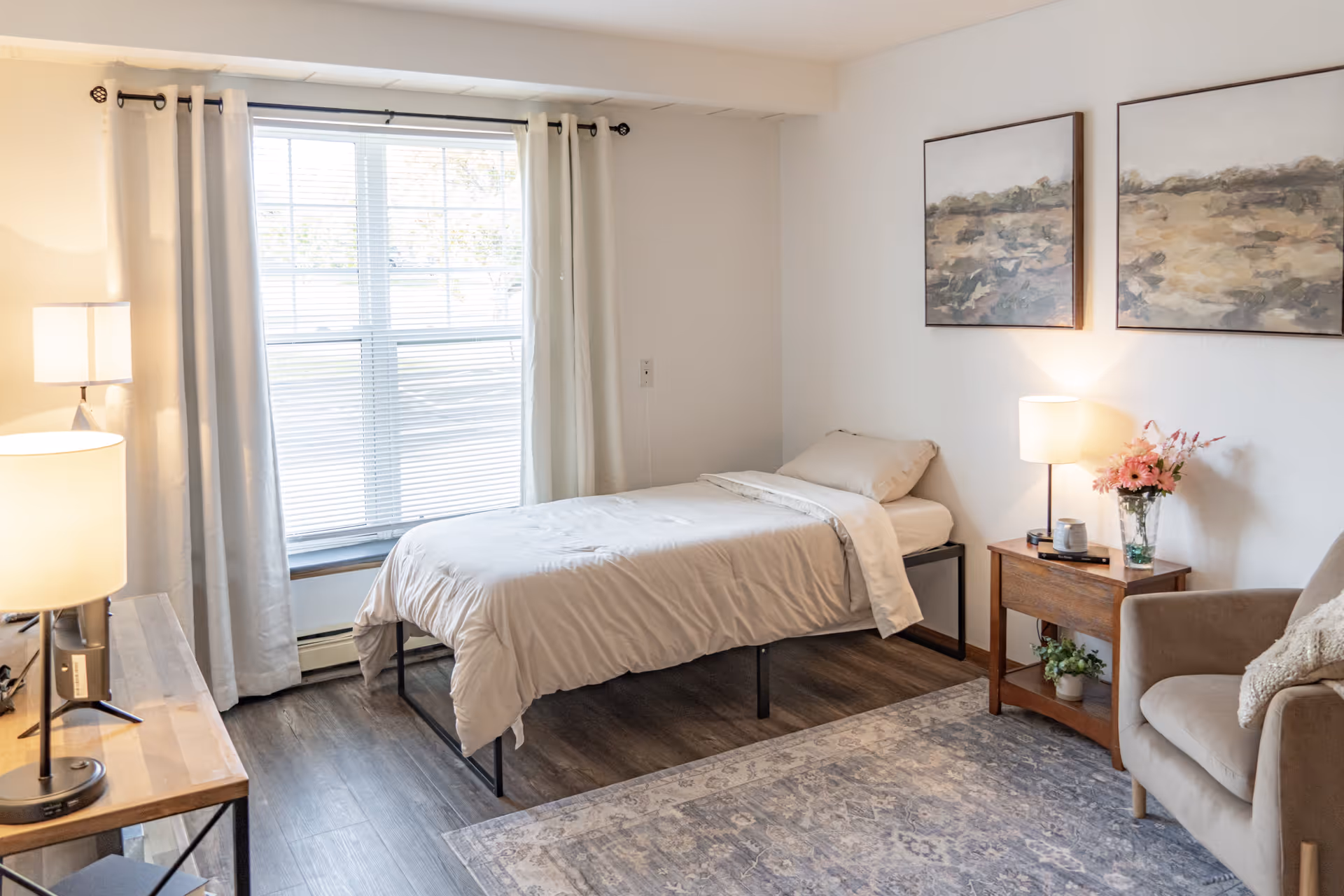 A bright and cozy bedroom with a single bed covered in beige bedding, a wooden nightstand with a lamp and a vase of flowers, a comfortable armchair with a cushion, two framed landscape paintings on the wall, a large window with white curtains, and a wooden desk with a lamp.