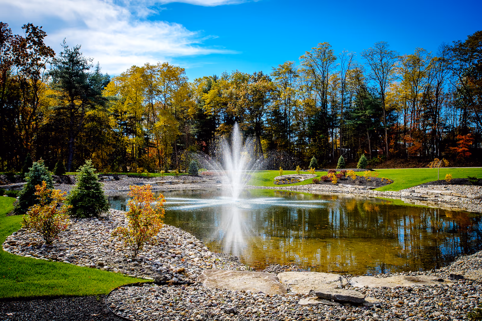 A landscaped outdoor area featuring a pond with a central water fountain surrounded by rocks, small shrubs, and trees with autumn foliage under a partly cloudy blue sky.