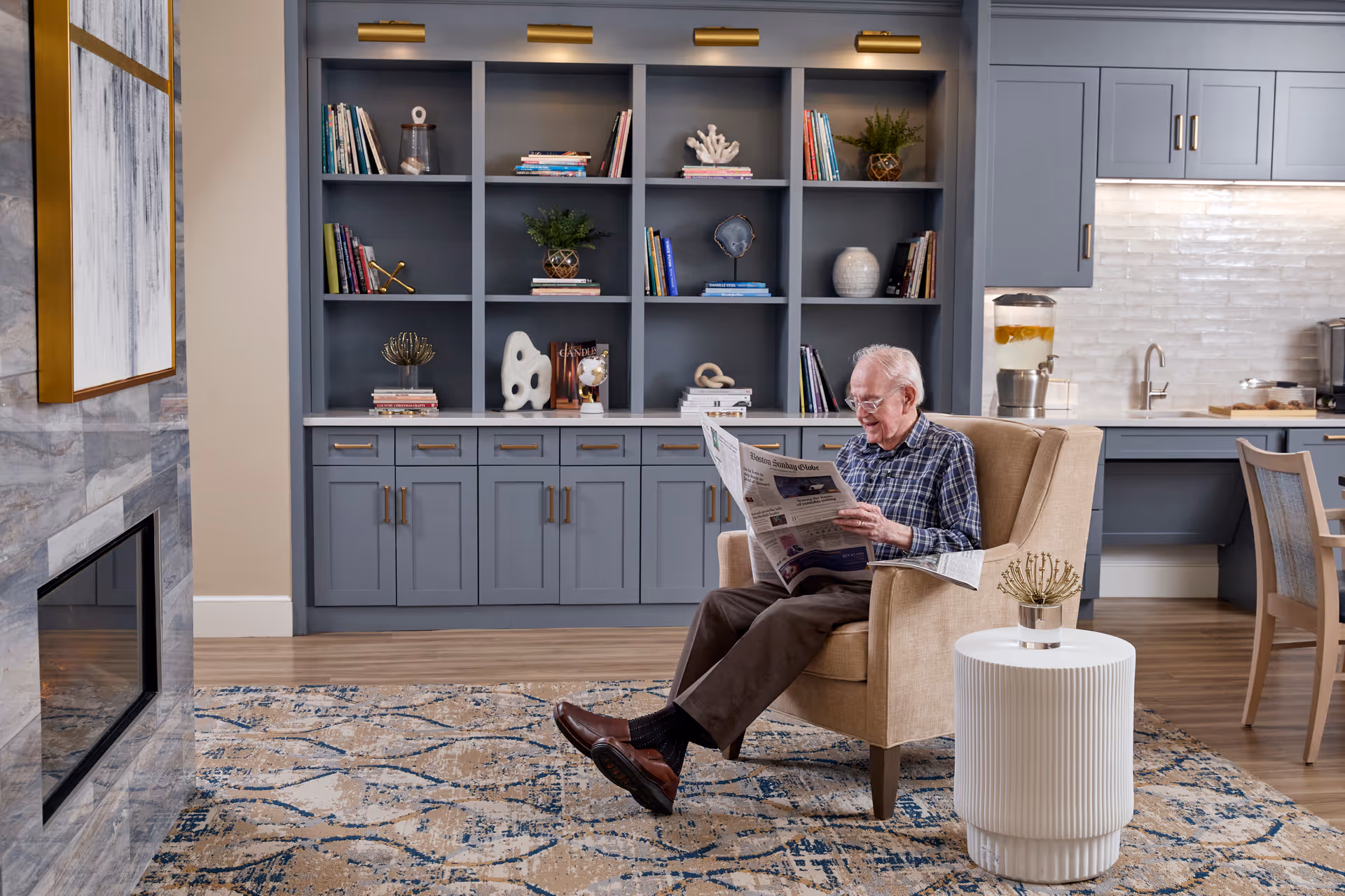 An elderly man sitting in a beige armchair reading a newspaper in a cozy living area. Behind him is a built-in blue-gray bookshelf with various books and decorative items. To the right, there is a kitchen area with cabinets, a sink, and a beverage dispenser. A patterned rug covers the wooden floor, and a small white side table with a decorative item is next to the armchair.
