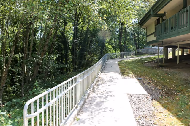 A paved outdoor walkway with a metal railing on the left side, surrounded by trees and greenery. On the right side, there is a building elevated on stilts with a green balcony. Sunlight filters through the trees, casting shadows on the path.