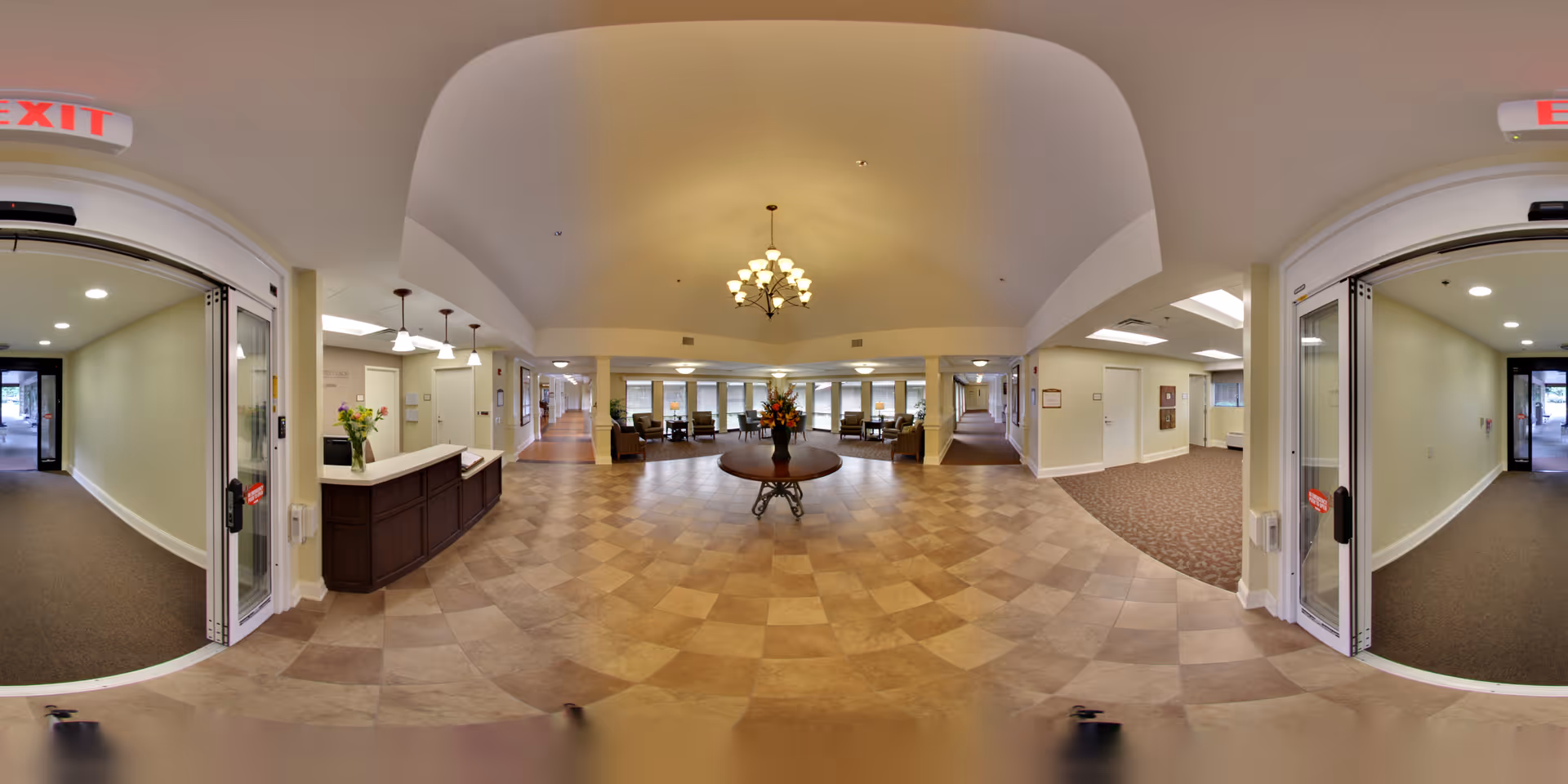 Wide-angle view of a senior living facility lobby with a reception desk, central table with flowers, seating area, and automatic double doors.