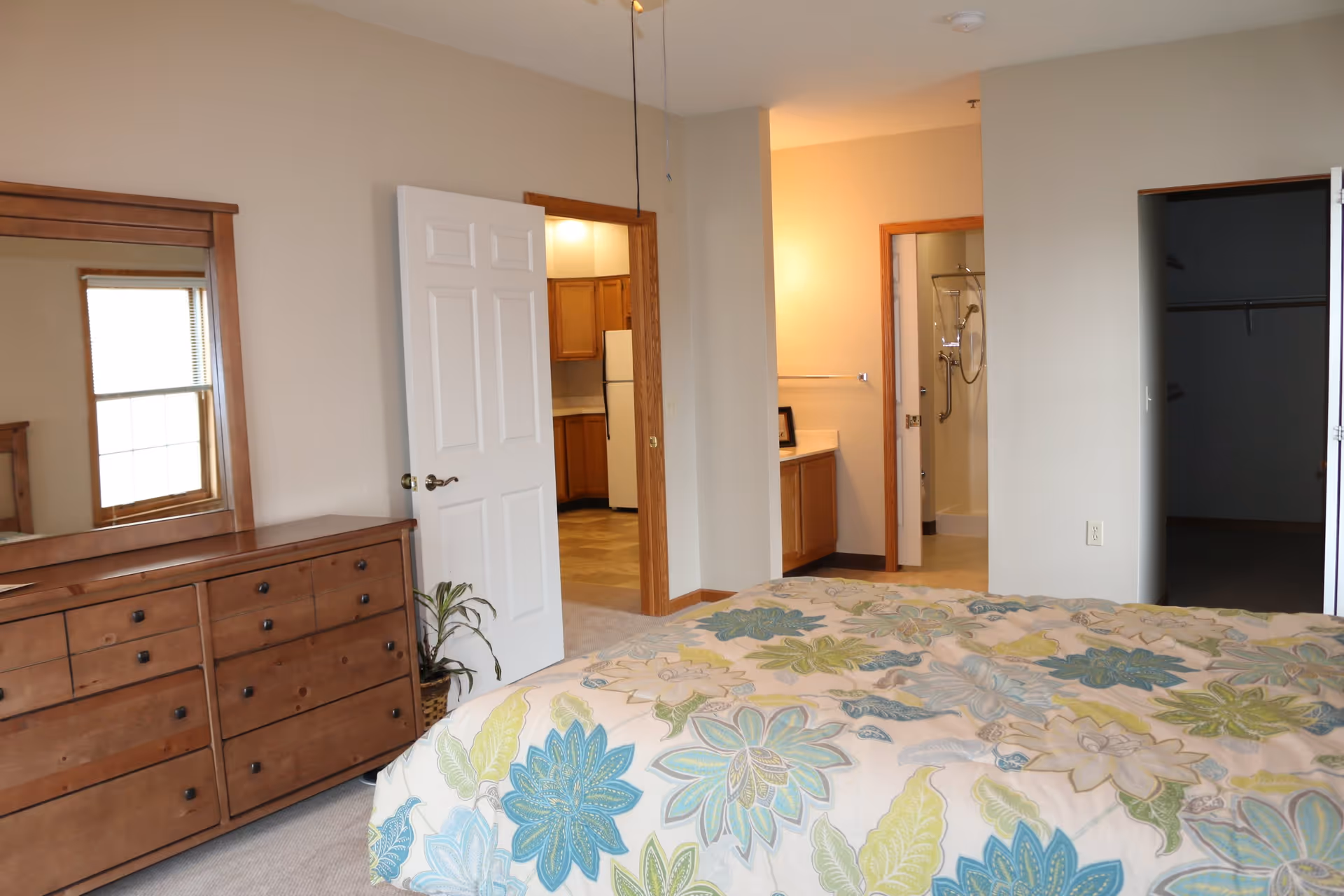 A bedroom with a bed covered in a floral patterned comforter in shades of blue, green, and beige. There is a wooden dresser with a large mirror on the left side, a window with blinds reflected in the mirror, and an open door leading to a kitchen area with wooden cabinets and a white refrigerator. Another open door reveals a bathroom with a shower and a vanity with a countertop and a towel rack. There is also an open closet space on the right side of the image.
