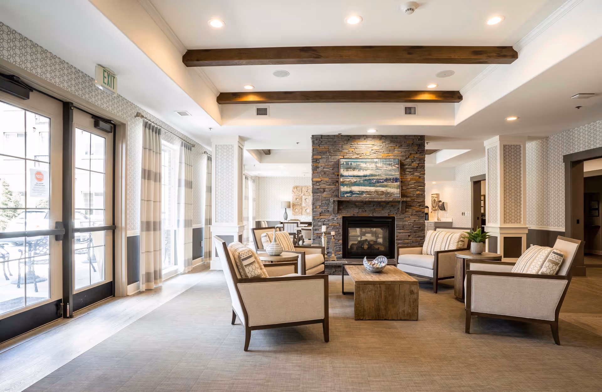 A bright and spacious living room area in an assisted living facility featuring a stone fireplace with a painting above it, surrounded by four beige armchairs with patterned cushions and a wooden coffee table. Large windows with sheer curtains allow natural light to fill the room, and wooden ceiling beams add a warm touch to the modern decor.