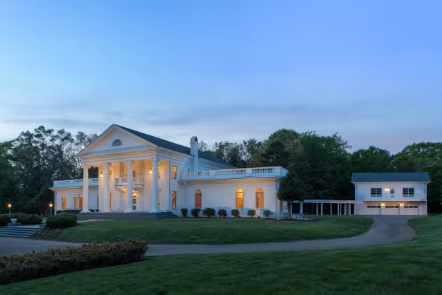 A large white colonial-style building with tall columns and warm interior lights glowing through the windows at dusk, surrounded by green lawns and trees. There is a driveway leading to a detached garage or secondary building on the right.