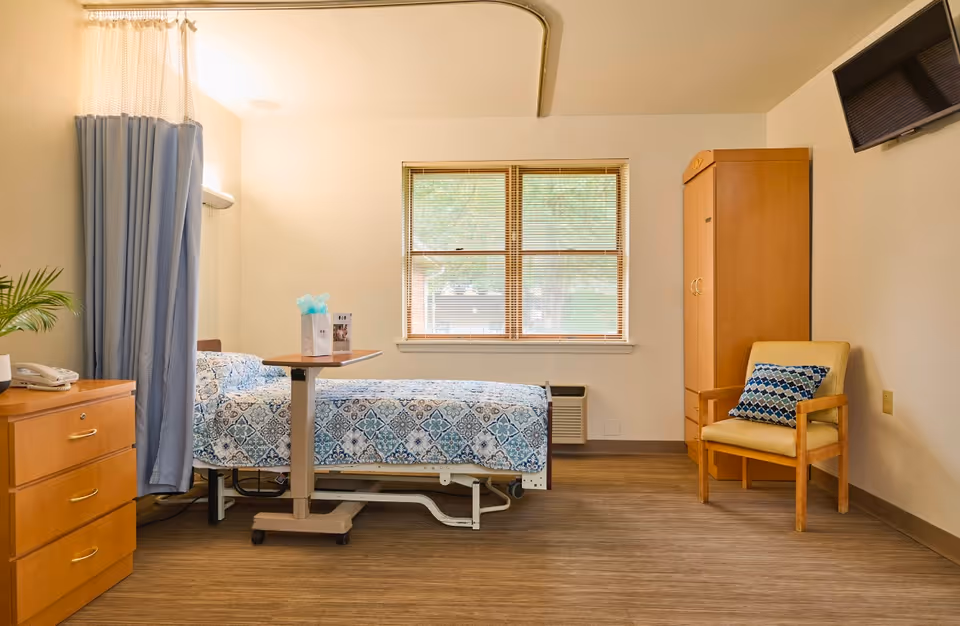 A bright senior care bedroom with a single bed, privacy curtain, overbed table, wooden dresser, wardrobe and an armchair beneath a wall-mounted TV.