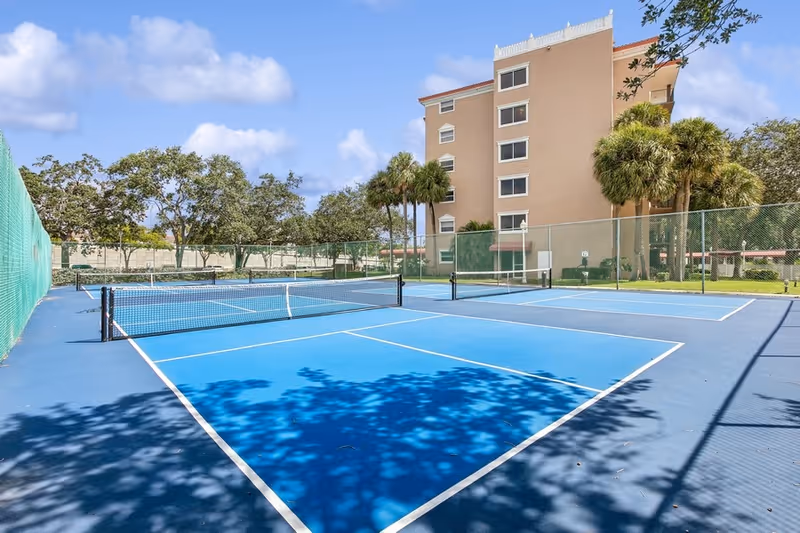 Outdoor tennis court with blue playing surface and white lines, surrounded by green fencing. In the background, there is a multi-story beige building and several palm trees under a partly cloudy blue sky.
