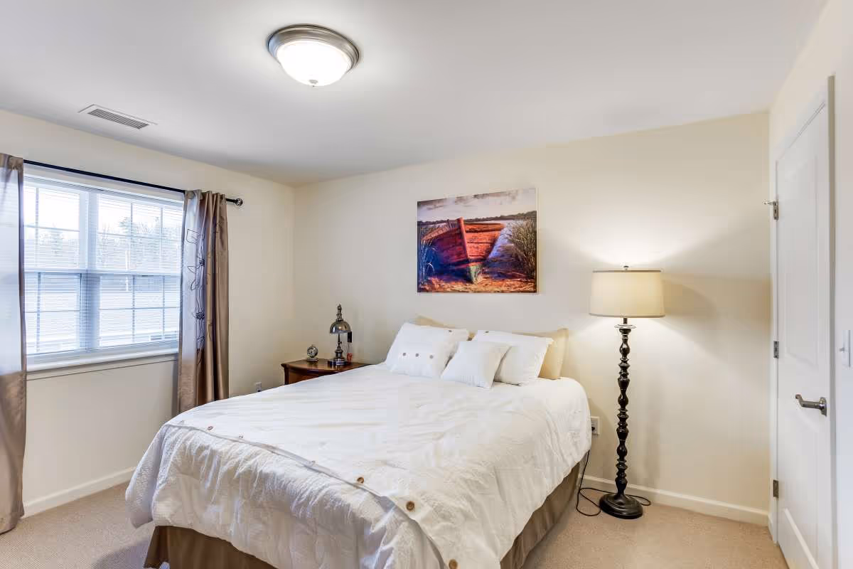 A bedroom with a large bed covered in white bedding and three pillows. There is a wooden nightstand with a lamp and clock on the left side of the bed, and a tall floor lamp on the right side. A window with beige curtains is on the left wall, and a painting of a red boat on water hangs above the bed. The walls are light-colored and the room has a beige carpet.