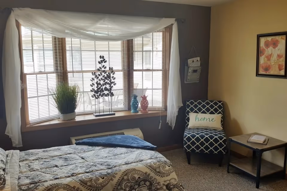 A cozy bedroom with a bed covered in a patterned quilt, a large window with blinds and white curtains, a decorative plant and figurines on the windowsill, a blue patterned chair with a pillow that says 'home', a small wooden side table with a book, and a framed floral picture on the wall.