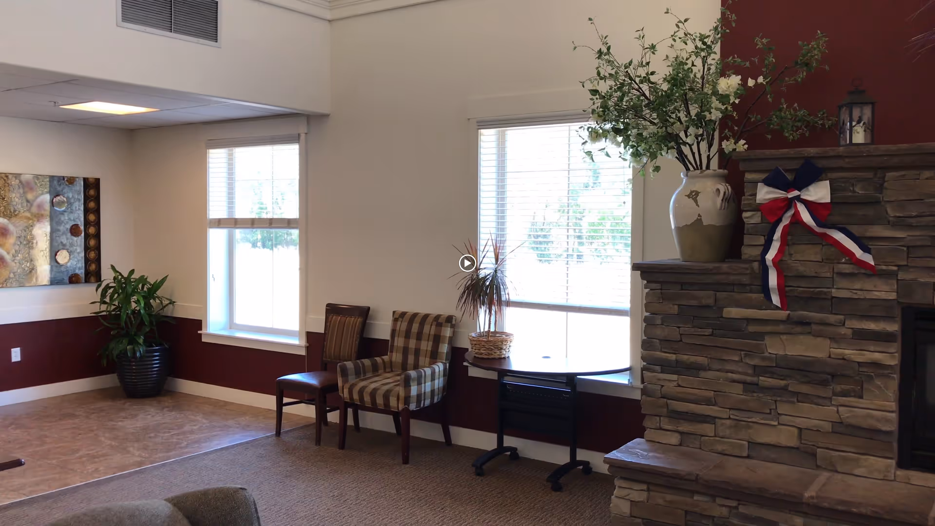 A cozy living room area in Tradition Assisted Living featuring a stone fireplace decorated with a large vase of flowers and a red, white, and blue ribbon. Two windows with blinds allow natural light into the room. There are two chairs, one with a plaid pattern and one solid, placed near a small table. A potted plant and a piece of abstract wall art are also visible.