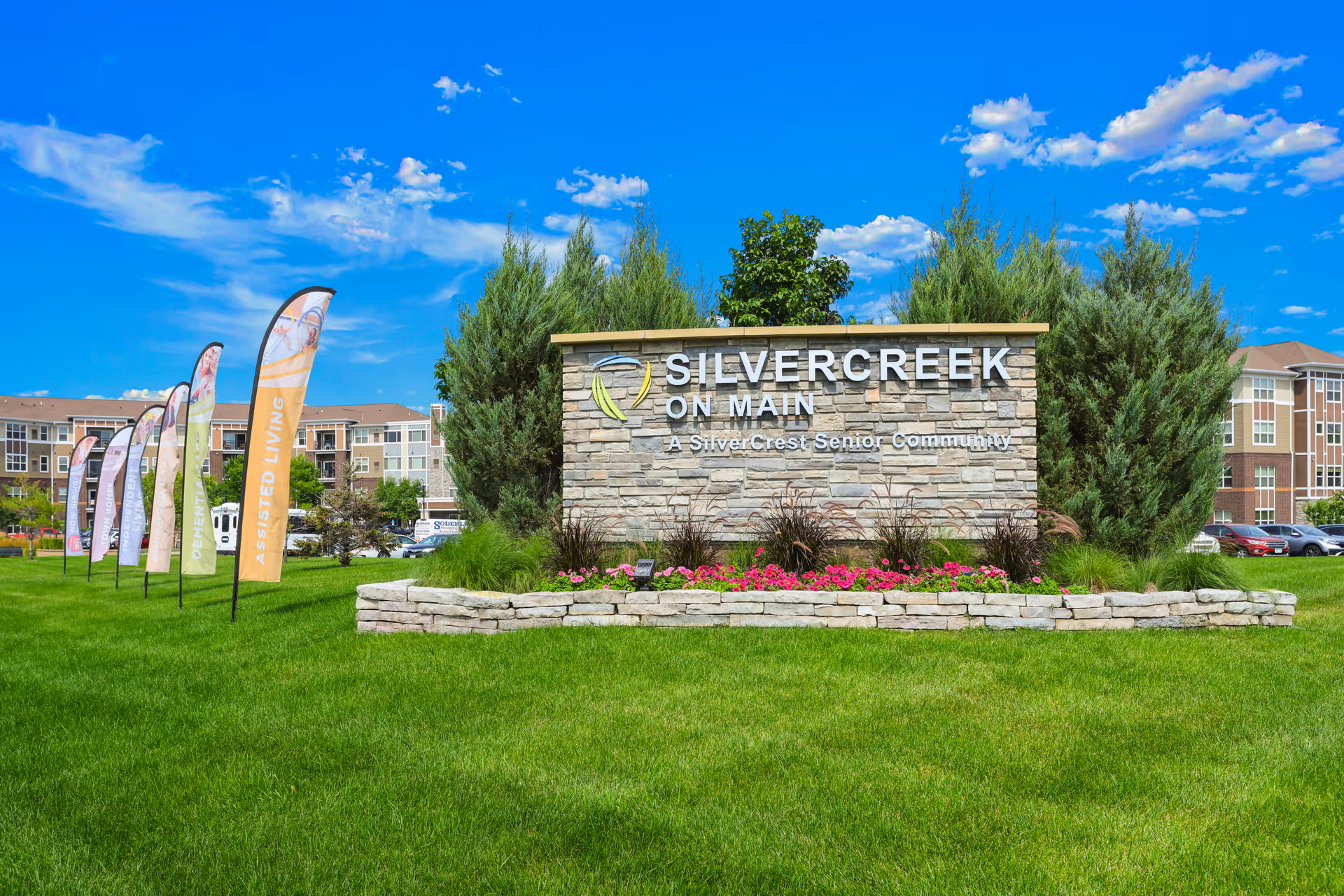 Stone sign for SilverCreek on Main, a SilverCrest Senior Community, surrounded by green grass, flowers, and trees with a bright blue sky and several colorful flags in the background.