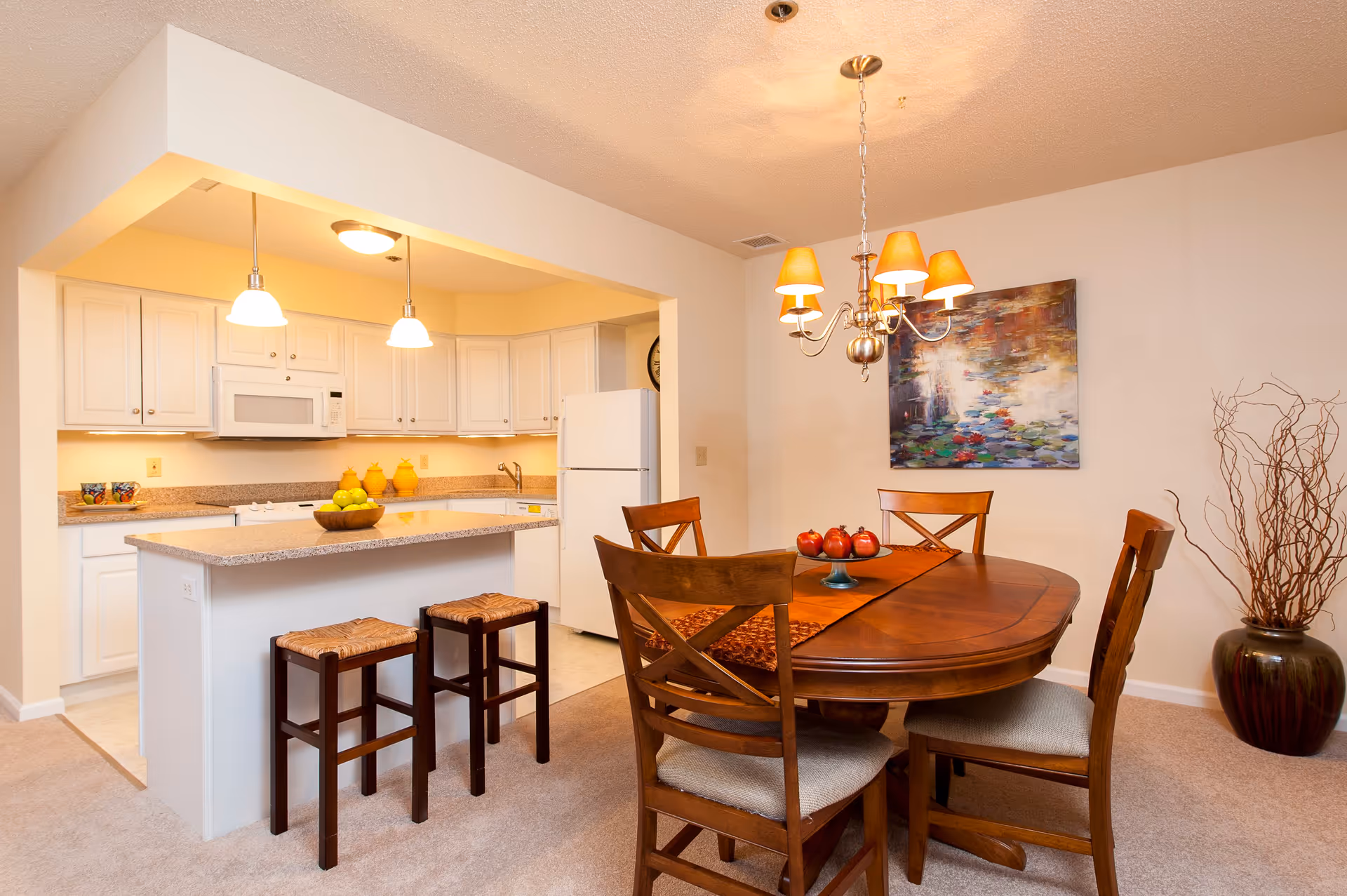 A cozy dining area with a wooden oval table and four chairs, decorated with a table runner and a centerpiece of red decorative items. Adjacent to the dining area is a kitchen with white cabinets, a white refrigerator, a microwave, and a countertop with two wooden stools. The room is warmly lit by a chandelier above the table and pendant lights over the kitchen counter. A colorful painting hangs on the wall, and a large vase with decorative branches is placed in the corner.