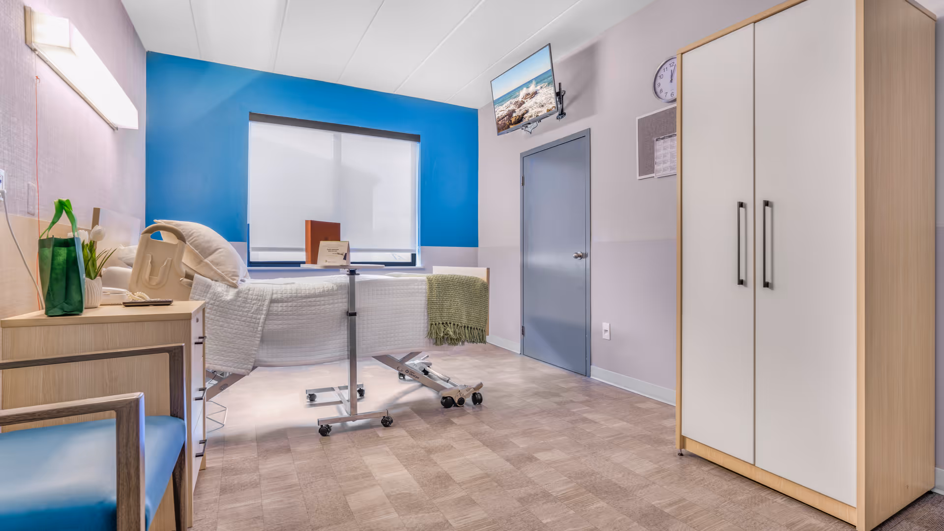 A clean and bright rehabilitation room with a hospital bed, a blue chair, a wooden cabinet, a wardrobe, a wall-mounted TV, and a window with a blue accent wall behind it.