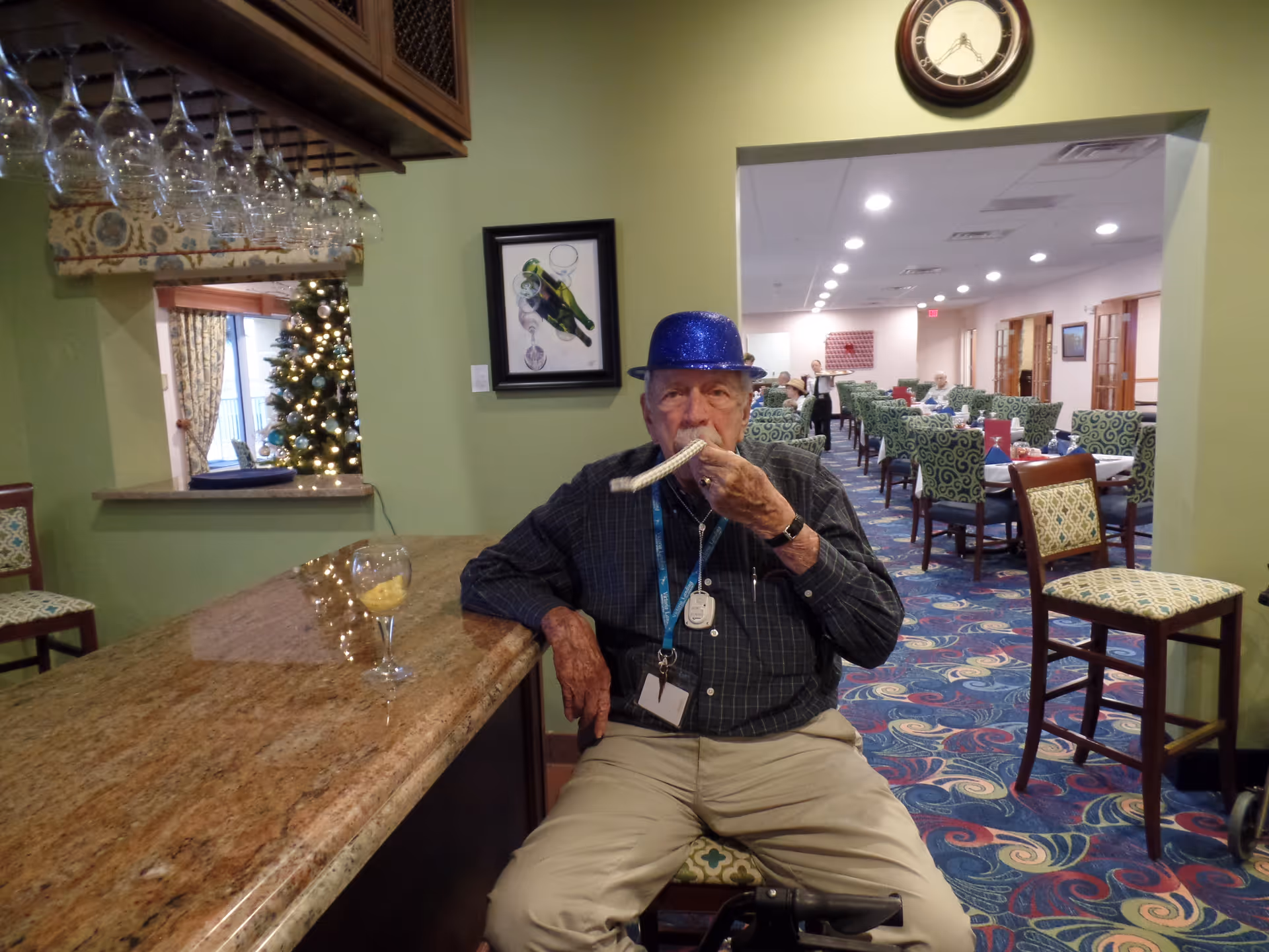 An elderly man wearing a blue glittery hat is sitting on a chair at a bar counter inside a senior living facility. He is blowing a party horn. Behind him is a dining area with tables and chairs, and a Christmas tree is visible through a window-like opening in the wall. The room has green walls and a colorful carpet.