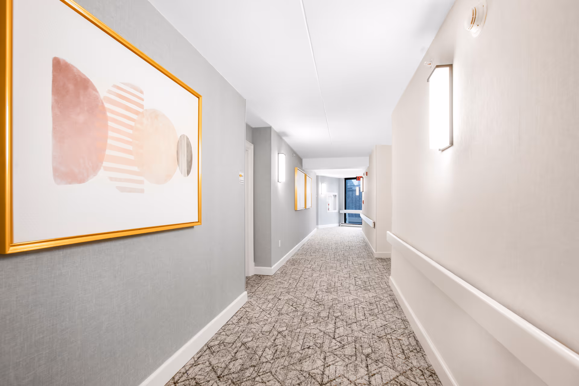 A well-lit, clean hallway in a senior living facility with patterned carpet, light gray walls, handrails on both sides, modern wall sconces, and abstract artwork framed in gold on the left wall. The hallway extends toward a door with a window at the far end.