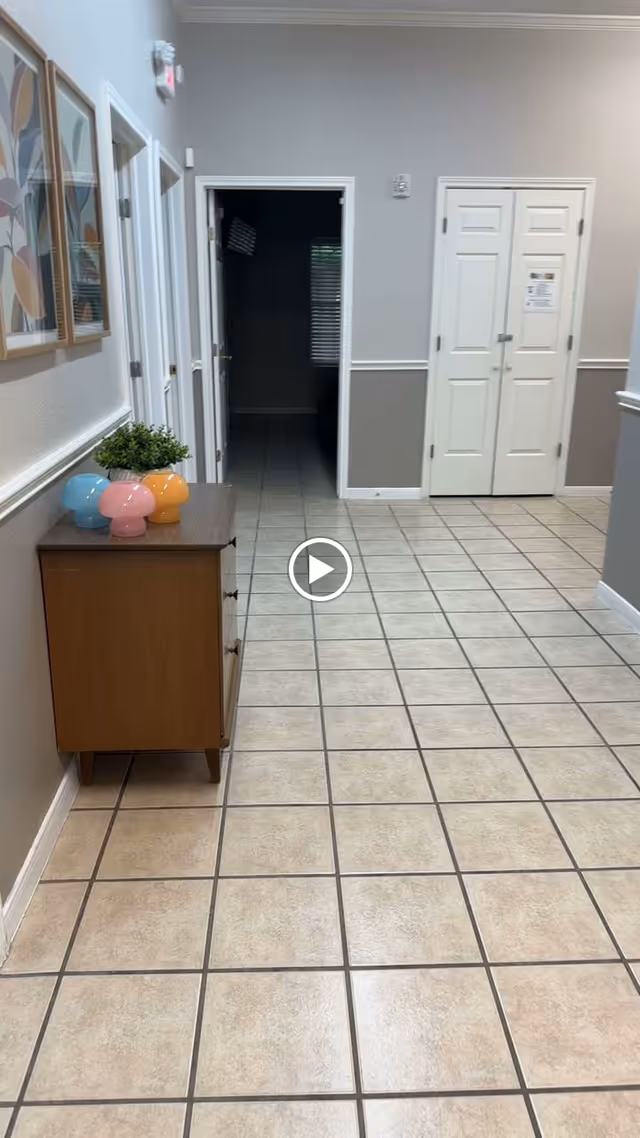Interior hallway with beige tiled floor and light gray walls. On the left side, there is a wooden dresser with three colorful mushroom-shaped decorations and a small green plant. Above the dresser, two framed abstract artworks hang on the wall. Several white doors are visible along the hallway, with one door open leading to a darker room. At the end of the hallway, there is a set of double white doors with a notice posted on one door.