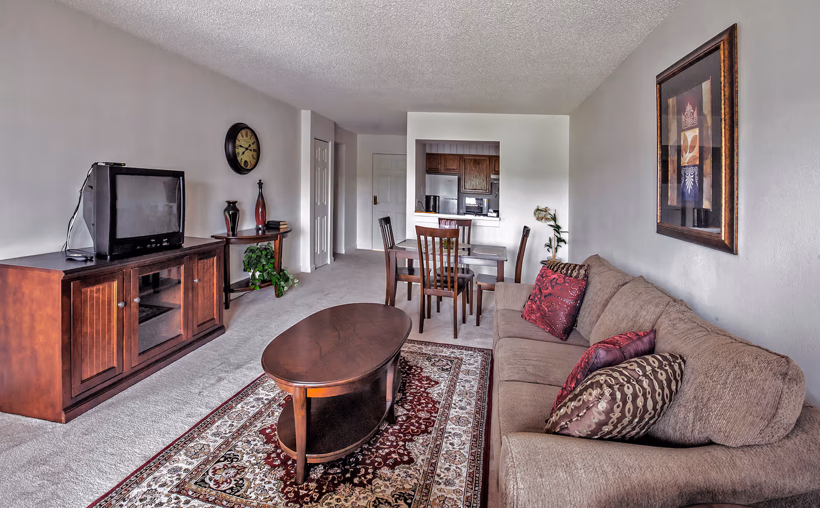 A living room area with a beige sofa adorned with patterned pillows, a wooden coffee table on a decorative rug, a wooden TV stand with an older style television, a wall clock, and a small table with decorative vases and a plant. In the background, there is a dining table with four chairs and a view into a kitchen area with wooden cabinets and appliances.