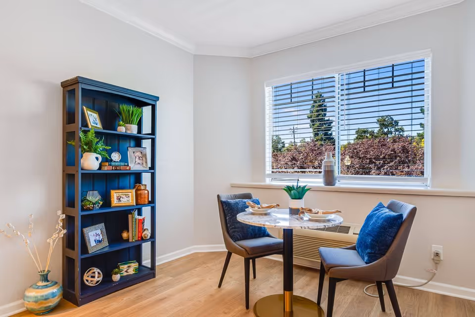 A cozy dining nook with a small round marble table set for two, two cushioned chairs with blue pillows, a large window with blinds showing trees outside, and a tall black bookshelf filled with decorative items and framed photos. A decorative vase with dried branches is placed on the floor beside the bookshelf.