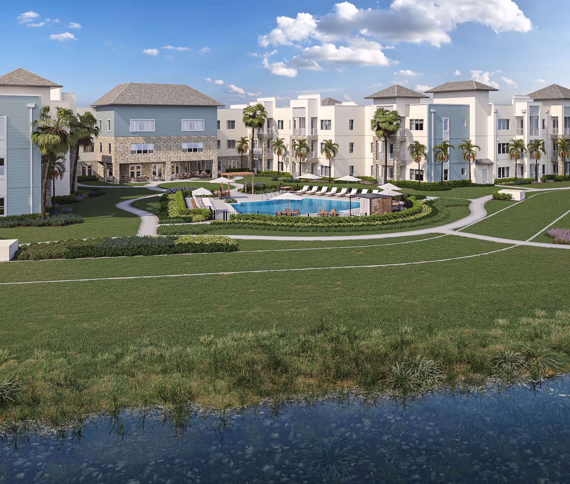 View of a senior living facility with multiple three-story buildings surrounding a central outdoor pool area. The pool is surrounded by lounge chairs, umbrellas, and greenery. Palm trees and well-maintained lawns are visible, with a small body of water in the foreground under a partly cloudy blue sky.