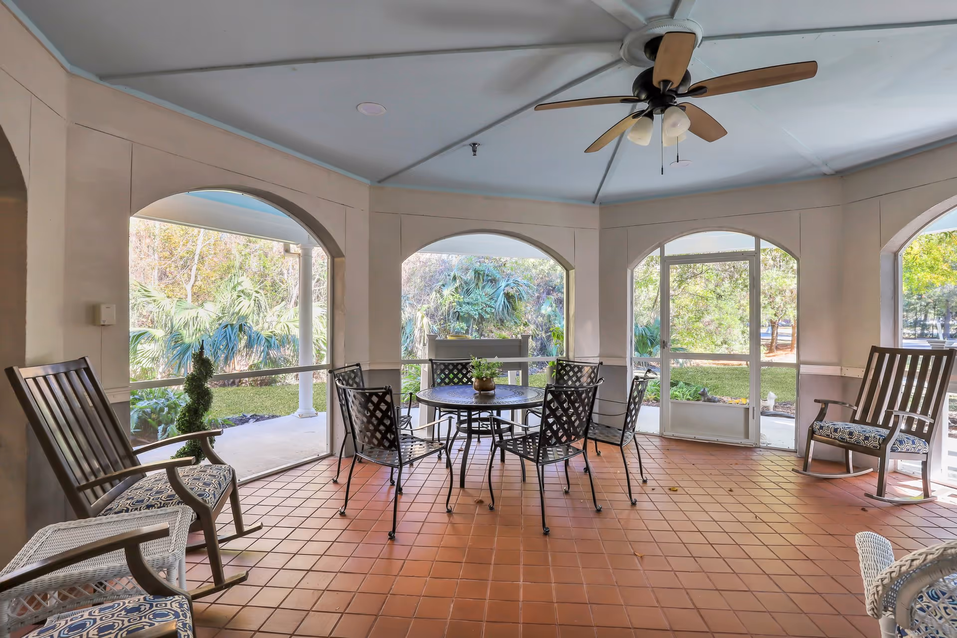 Screened-in covered patio with a round metal table and chairs, rocking chairs, a ceiling fan, and arched openings overlooking greenery.