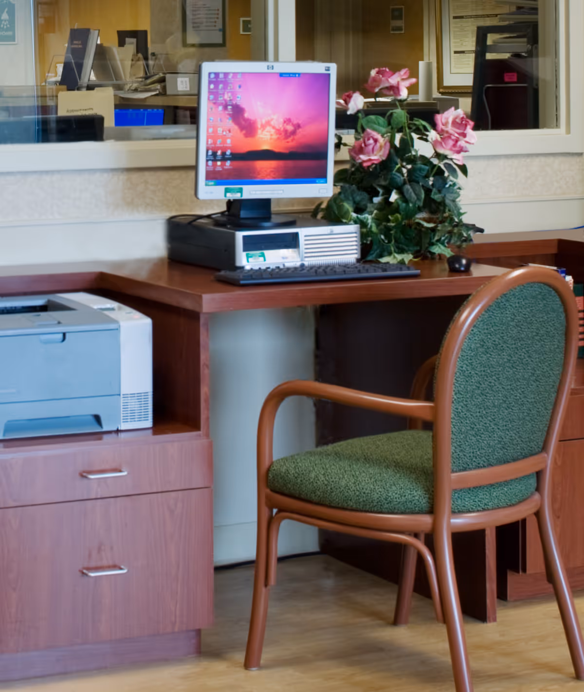 A wooden desk with a desktop computer, keyboard, mouse, and a potted plant with pink flowers. A green cushioned chair is positioned in front of the desk. There is a printer on a lower shelf to the left, and a window behind the desk showing an office area with papers and files.