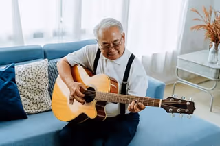 An elderly man sitting on a blue couch playing an acoustic guitar in a bright living room with white curtains and decorative pillows.