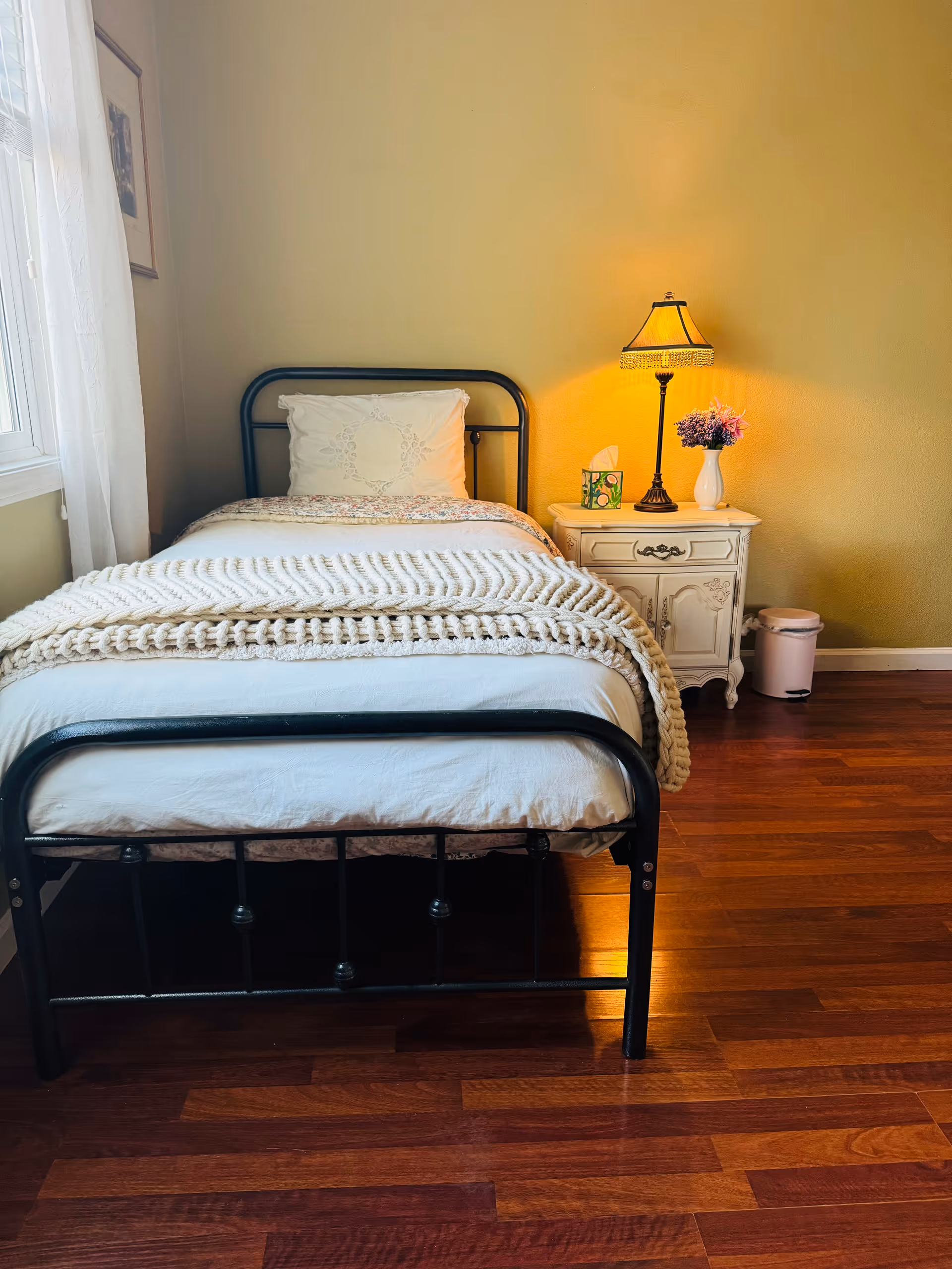 A cozy bedroom with a single black metal bed frame, white bedding, and a chunky knit blanket. Next to the bed is a white vintage-style nightstand with a table lamp, a box of tissues, and a small vase with purple flowers. The room has wooden flooring and light yellow walls, with a window covered by sheer white curtains on the left side.