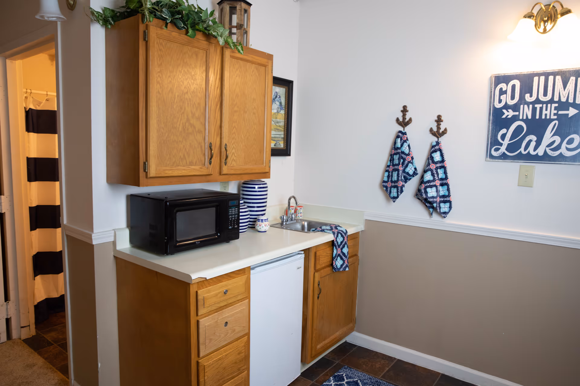 Small kitchenette with wooden cabinets, a microwave, sink, mini-fridge, decorative towels and a 'Go Jump in the Lake' sign on the wall.