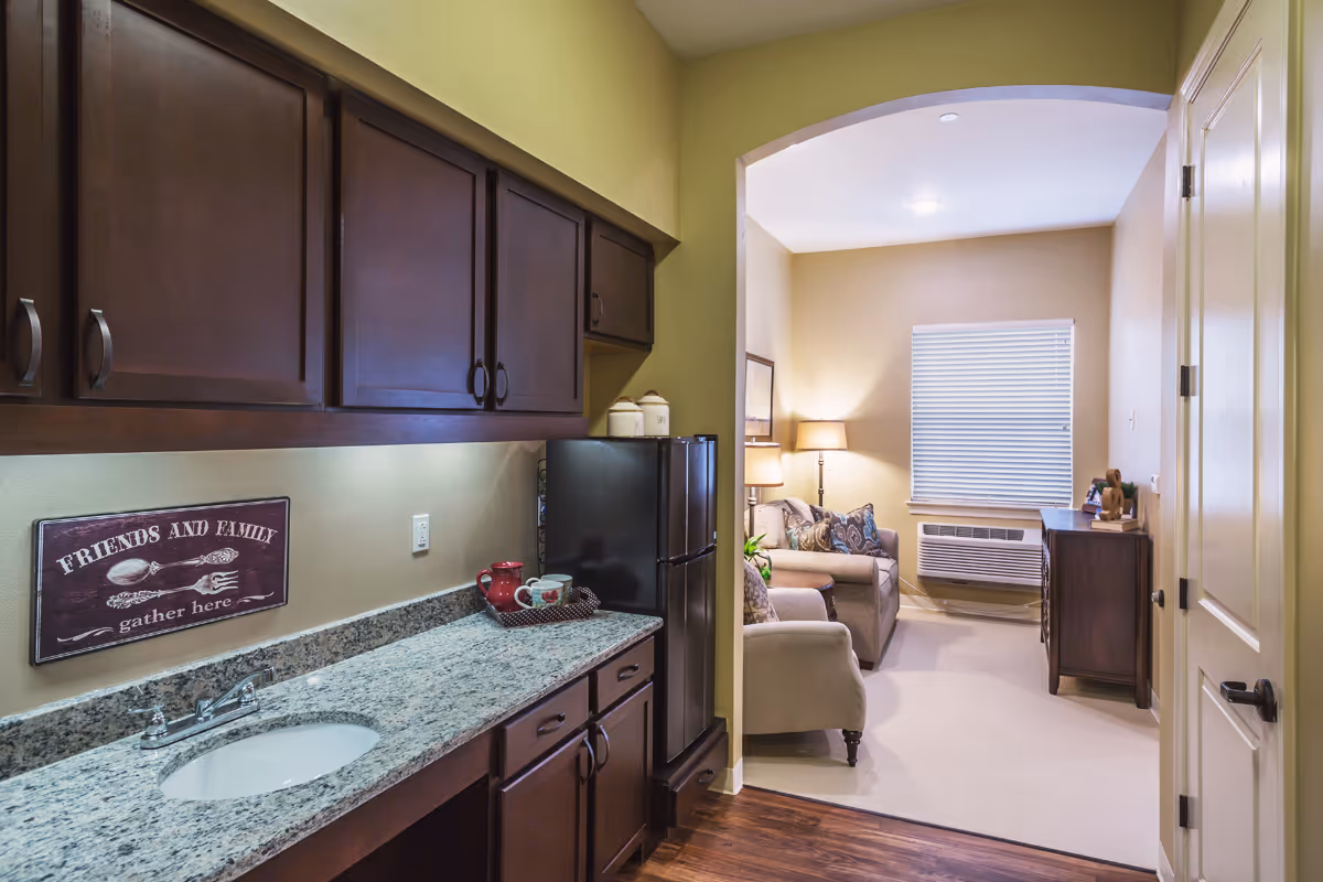 Small kitchenette with dark wood cabinets and granite countertop opening to a cozy living room with a sofa, lamp, and window air conditioner.