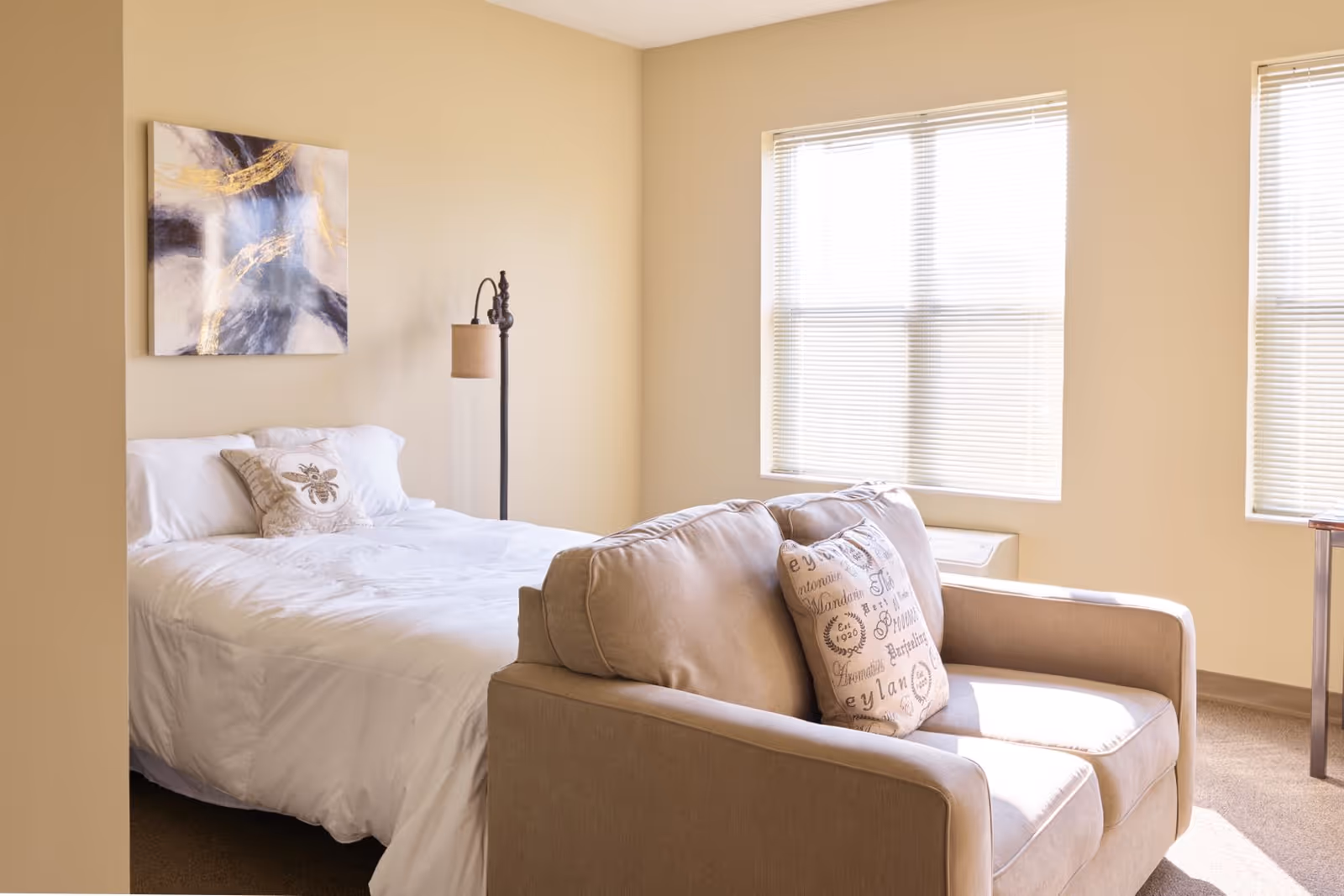 Sunlit bedroom with a made bed, a beige loveseat at the foot, decorative pillows, wall art, and two windows with blinds.