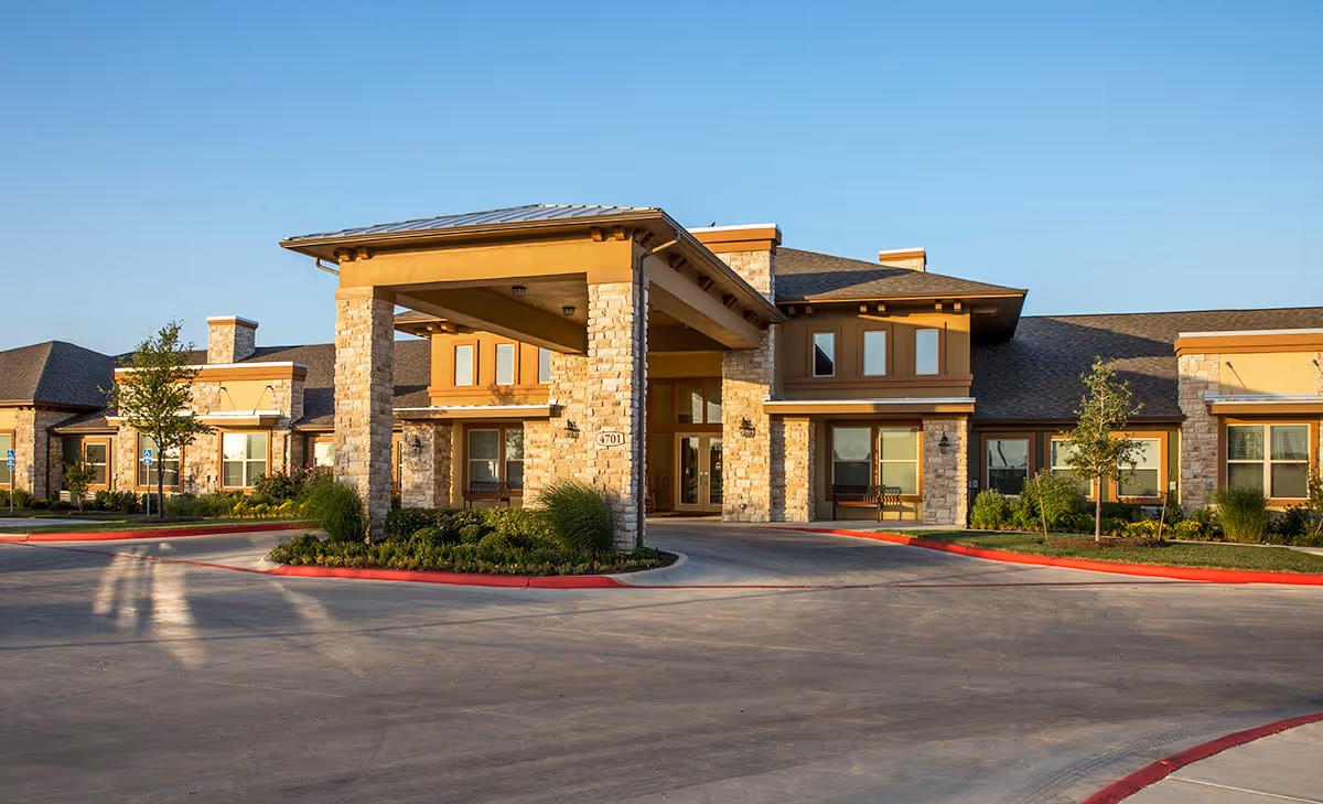 Front entrance of a single-story senior living building with a covered porte-cochère, stone facade, and landscaped circular driveway.