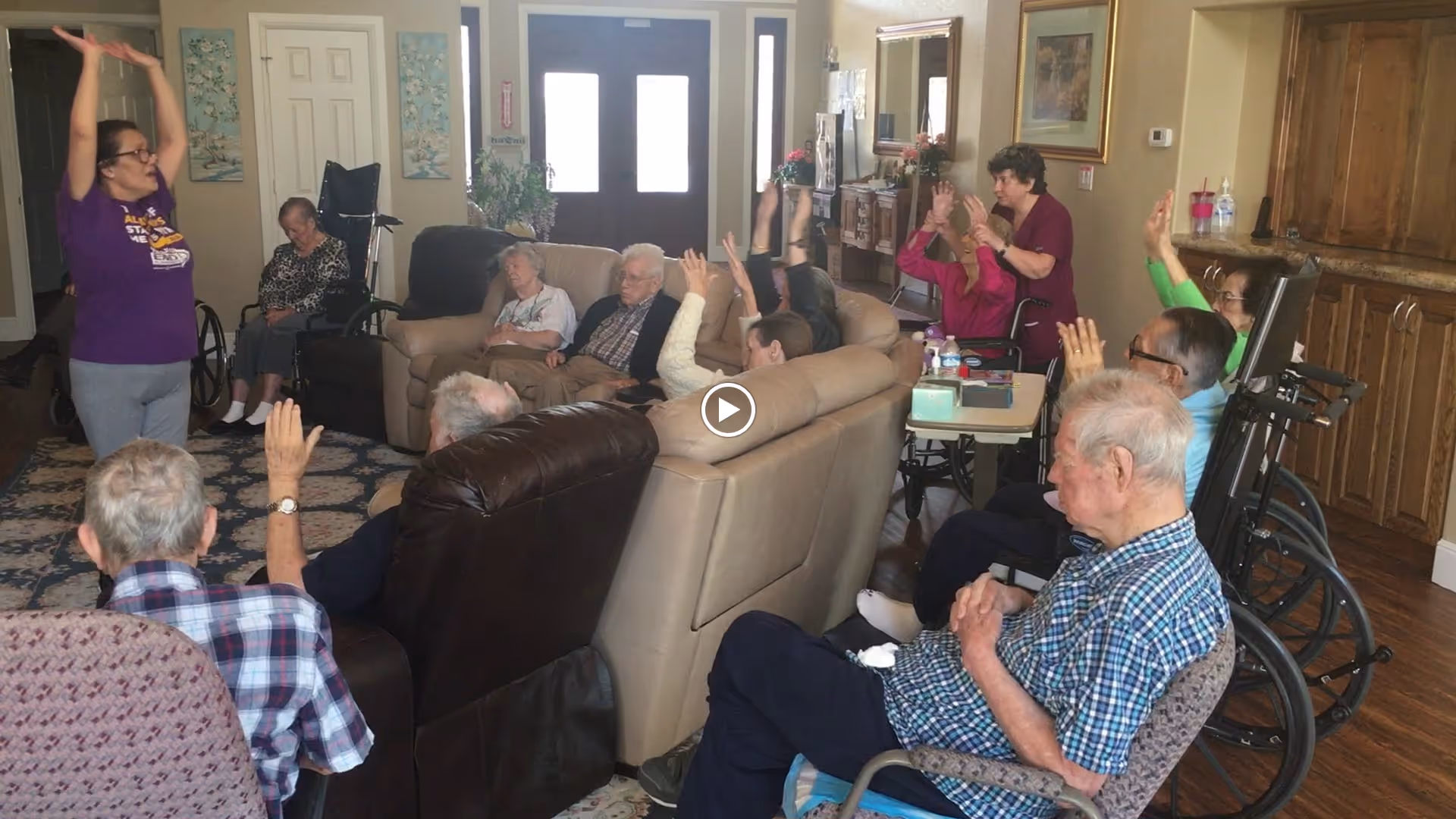 A group of elderly people seated in a living room area, some in wheelchairs, participating in a group activity led by a woman standing with her arms raised. The room has comfortable seating, wooden cabinets, and a carpeted floor.