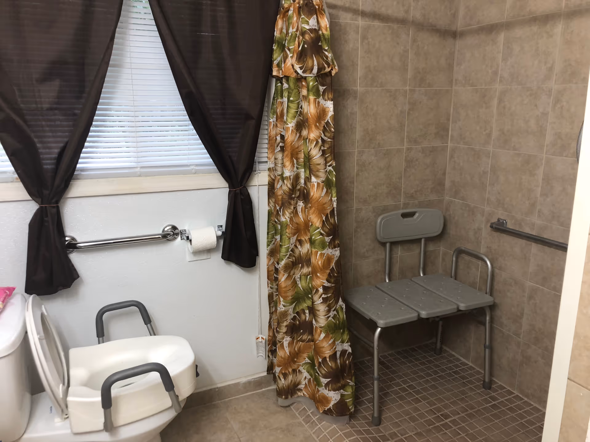 A bathroom with a toilet equipped with a raised seat and armrests, a grab bar on the wall with a toilet paper roll, a window with brown curtains, and a tiled shower area with a floral shower curtain and a gray shower chair with backrest and armrests.