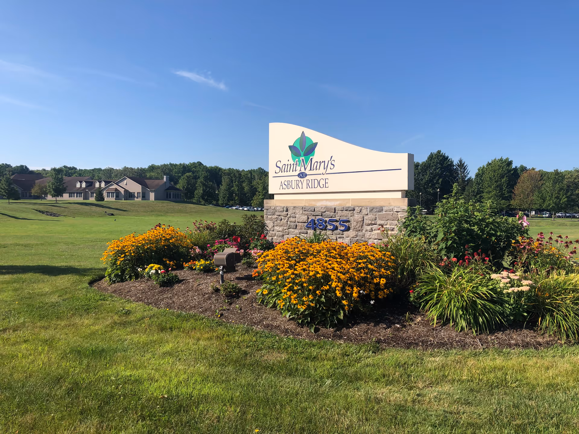 Outdoor view of the entrance sign for Saint Mary's at Asbury Ridge surrounded by colorful flowers and greenery, with a large building and trees in the background under a clear blue sky.