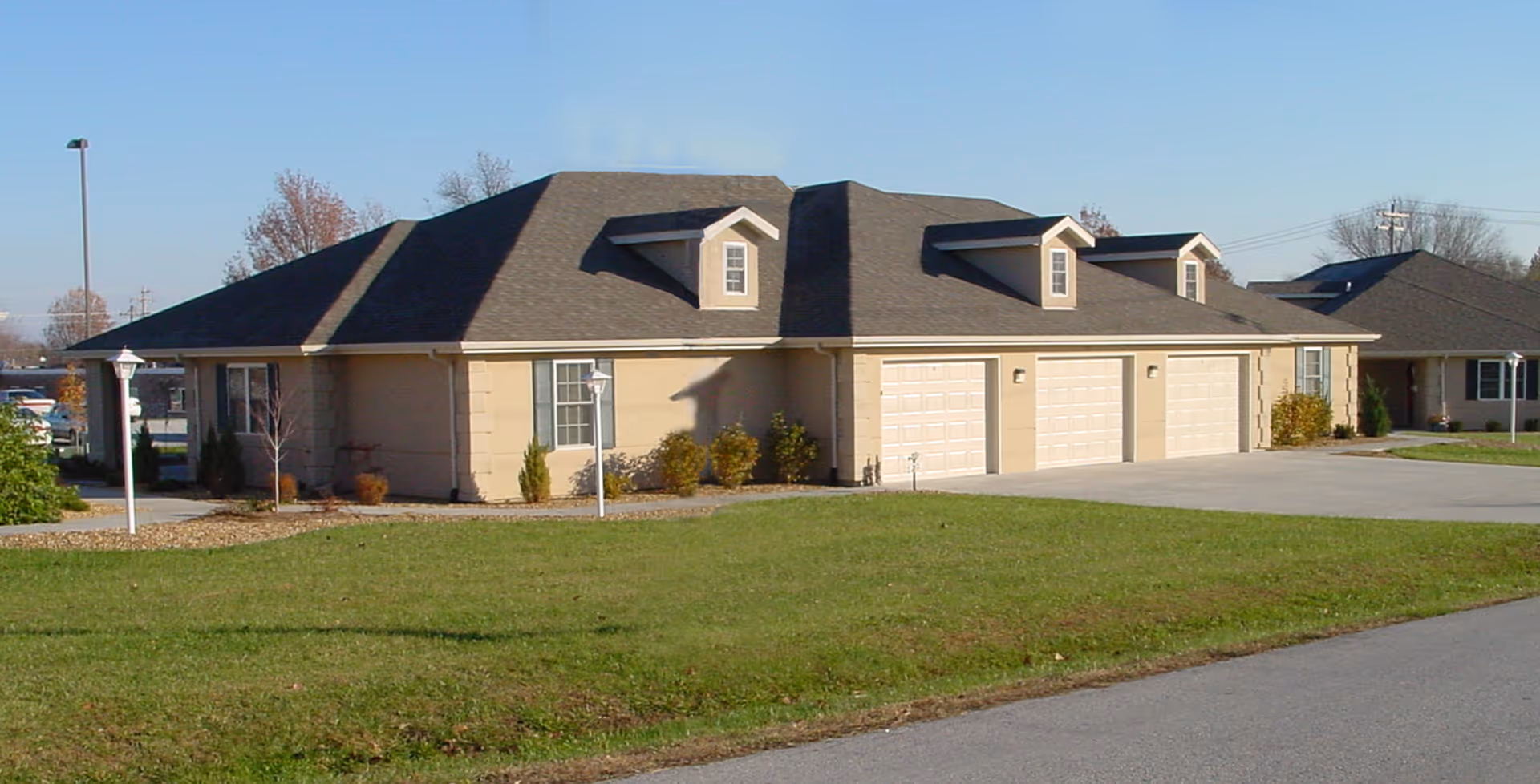 Exterior view of a single-story beige building with a dark shingled roof and three garage doors, surrounded by a green lawn and a paved driveway under a clear blue sky.
