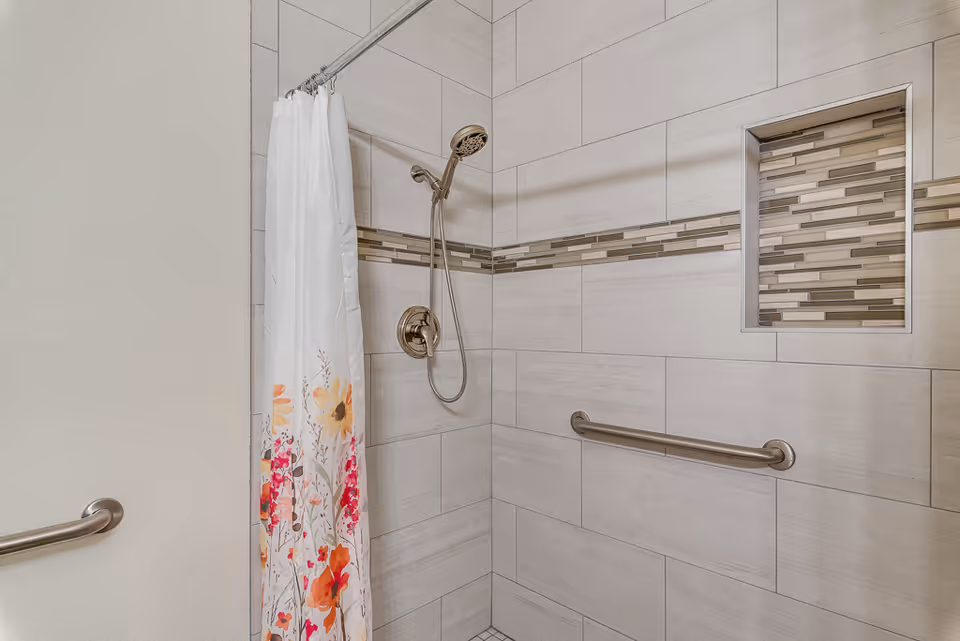 Tiled walk-in shower with a handheld showerhead, grab bars, a decorative tile niche, and a floral shower curtain.