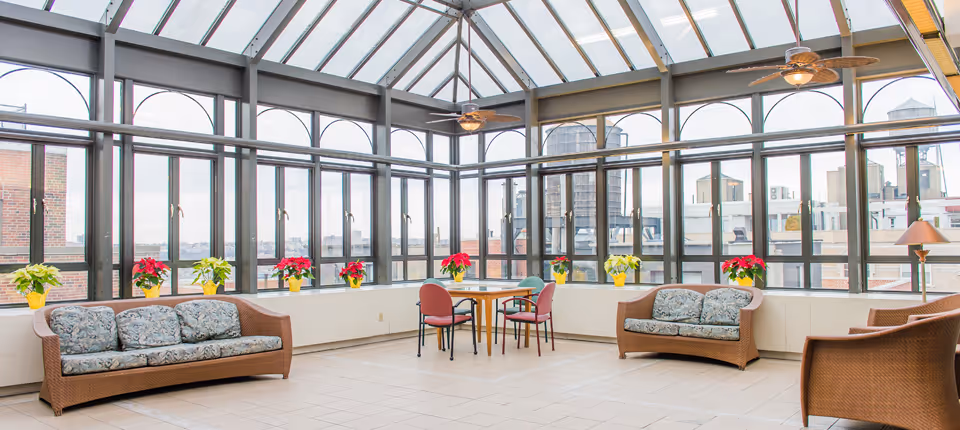 A bright indoor lounge area with large windows and a glass ceiling allowing natural light to fill the space. The room features two wicker sofas with floral cushions, a small round table with four chairs, several potted poinsettia plants on the windowsill, ceiling fans, and a floor lamp in the corner. Outside the windows, urban buildings and water towers are visible.