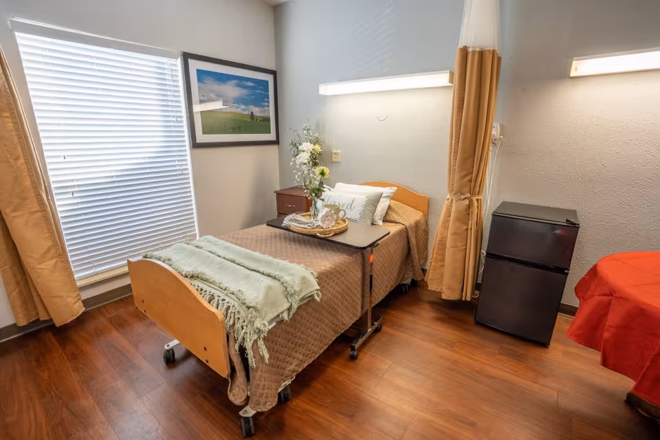 Bright senior living bedroom with a single bed and overbed tray, bedside table with flowers, mini refrigerator, window with blinds and curtains, and wood flooring.