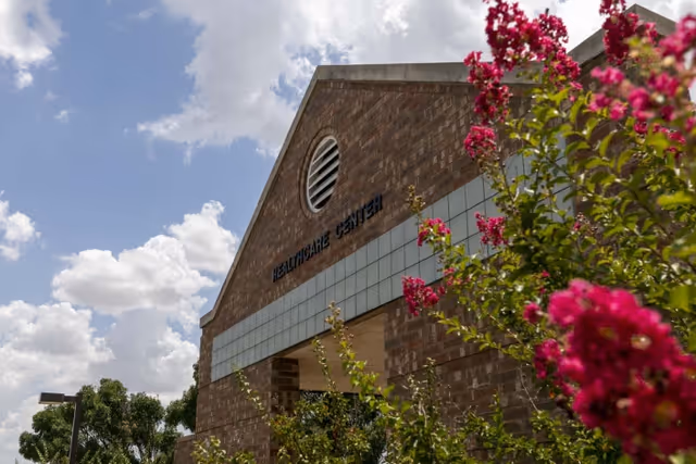 Brick healthcare center facade with pink flowering bushes in the foreground and a partly cloudy sky above.