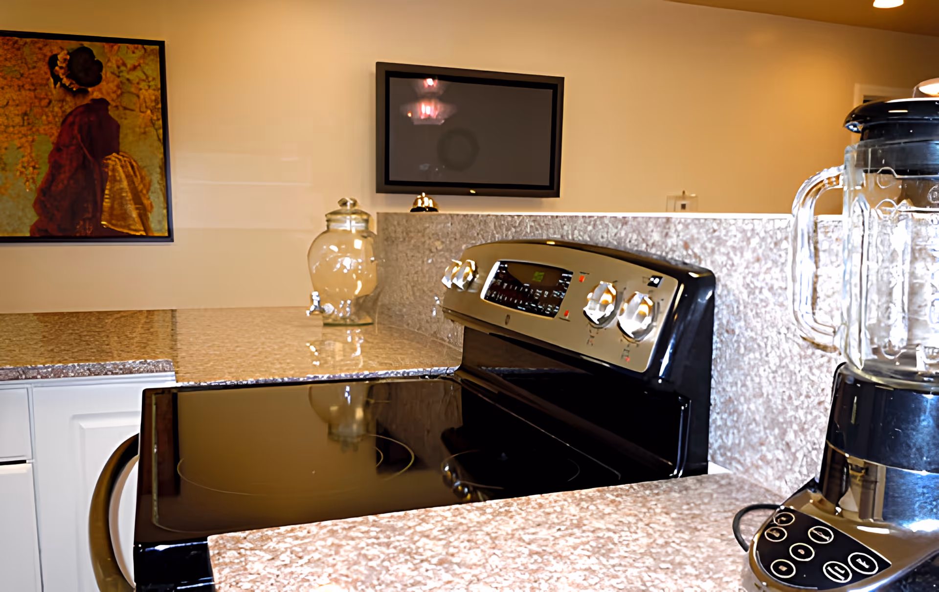 A modern kitchen countertop with a black electric stove, a blender, a glass jar, and a wall-mounted flat screen TV. There is a framed artwork on the wall to the left.
