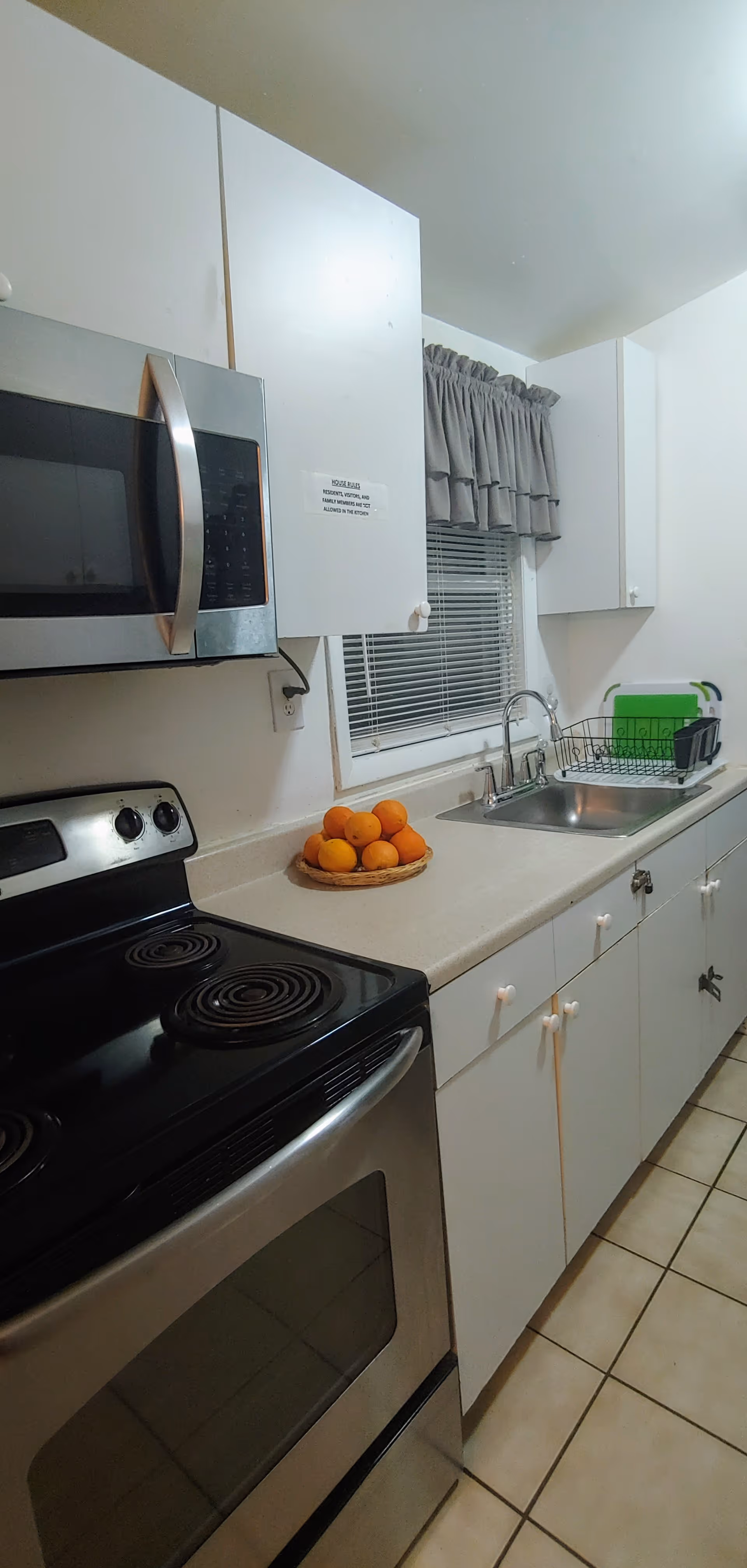 A small kitchen with white cabinets, a stainless steel electric stove and microwave, a countertop with a basket of oranges, a sink with a drying rack, and a window with gray curtains.