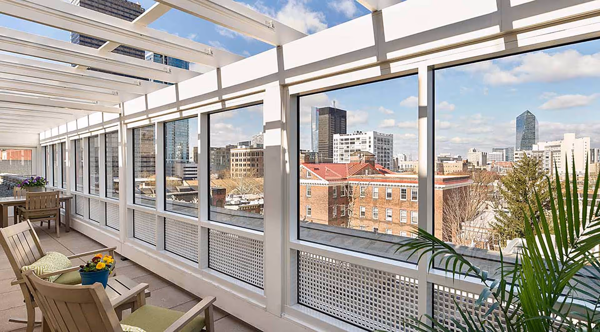 A bright enclosed balcony or sunroom with large windows offering a panoramic view of a city skyline. The space features wooden chairs with green cushions, a small table with a flower pot, and a potted plant in the corner. The ceiling has white beams allowing natural light to fill the area.