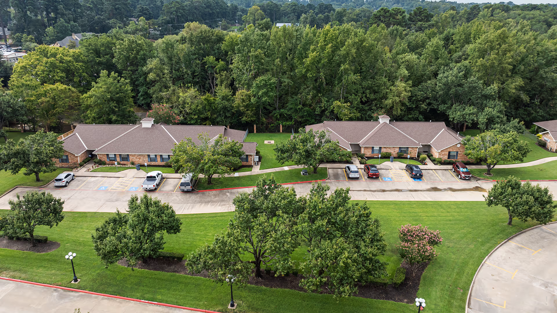 Aerial view of a senior living facility with two single-story brick buildings surrounded by green lawns, trees, and a parking lot with several cars parked. The background features a dense forested area.