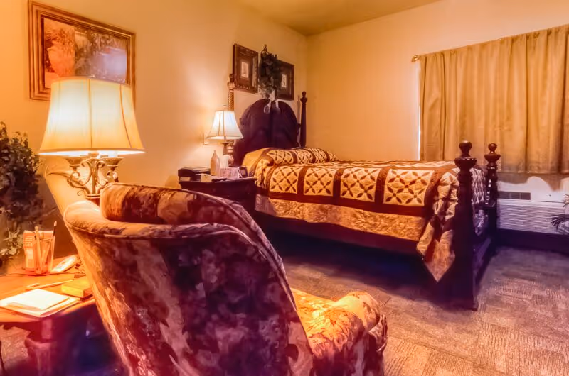 Cozy furnished bedroom with a wooden bed dressed in a patterned quilt, an upholstered armchair, bedside tables and lamps.