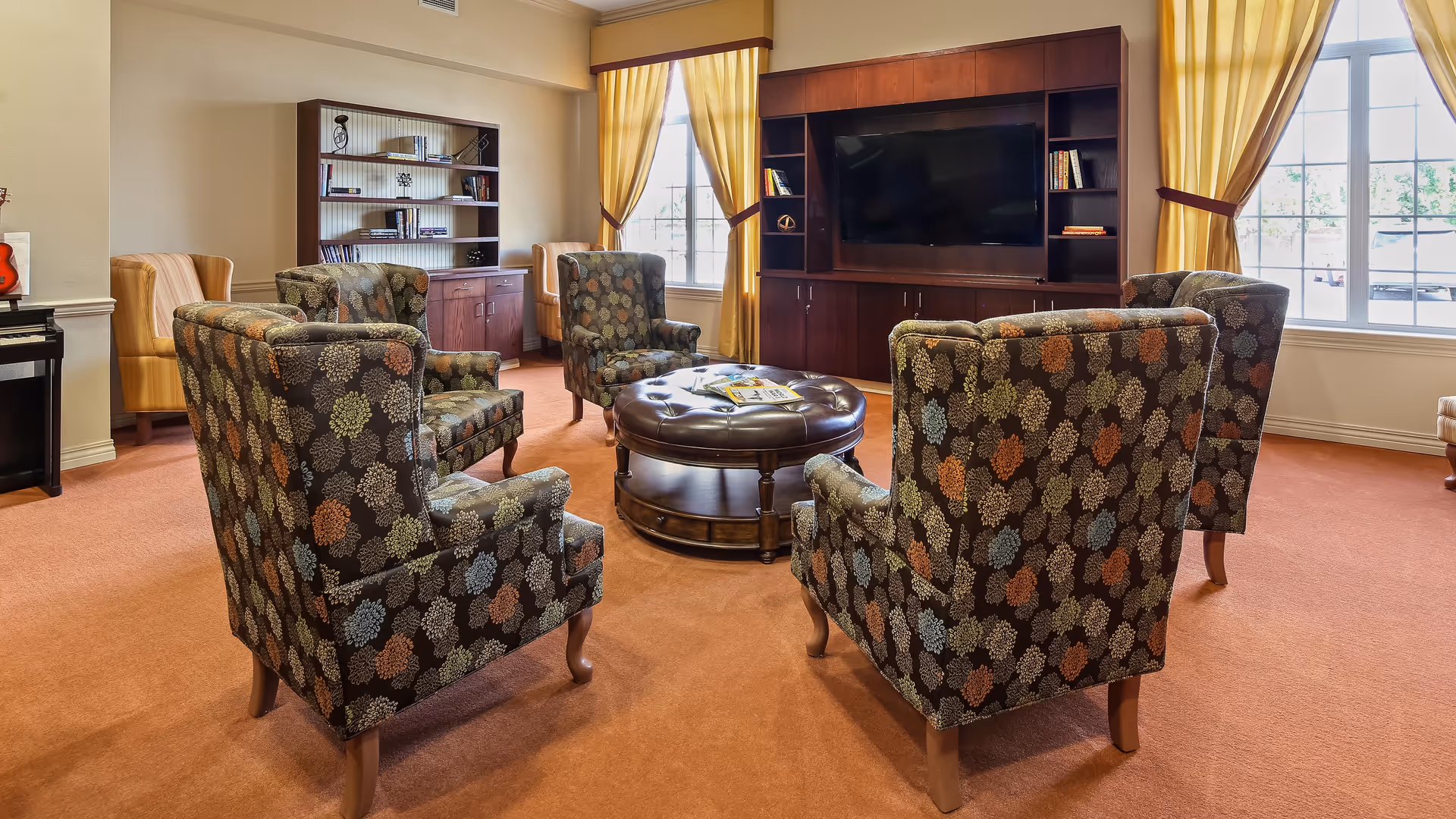 A cozy living room with four patterned armchairs arranged around a round leather ottoman. The room has large windows with yellow curtains, a wooden entertainment center with a flat-screen TV, and a bookshelf filled with books and decorative items. The carpet is a warm orange color.