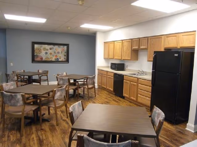 A communal dining area with several square tables and chairs arranged on a wooden floor. The room features a kitchenette along one wall with wooden cabinets, a black refrigerator, a microwave, a dishwasher, and a sink. A framed colorful artwork is hung on a blue accent wall.